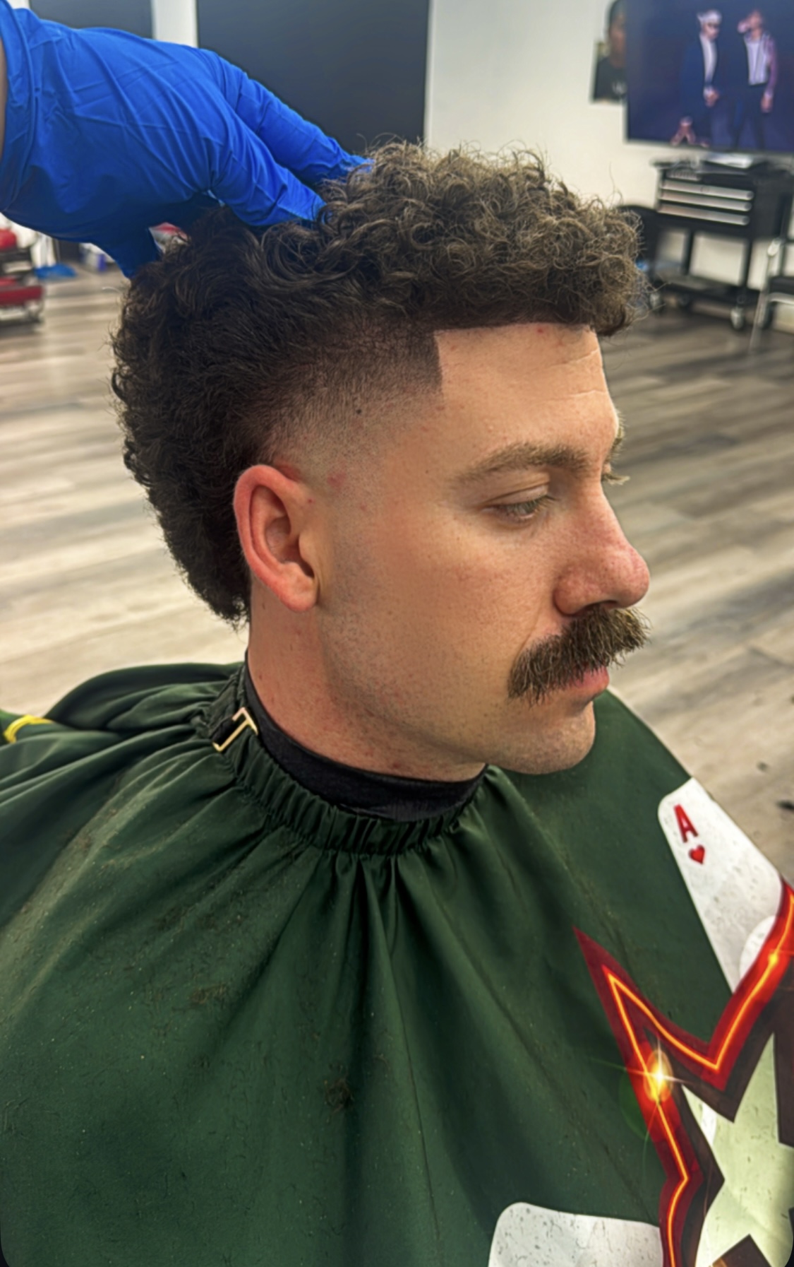 Man with a curly mullet, taper fade and mustache being styled by a gloved barber